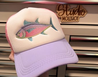 Puede incluir: Una gorra tipo trucker con panel frontal blanco y un bordado de un pez en tonos rosa, morado y turquesa. La gorra presenta una visera de color morado claro y una parte trasera de malla rosa, con una estructura frontal de espuma.