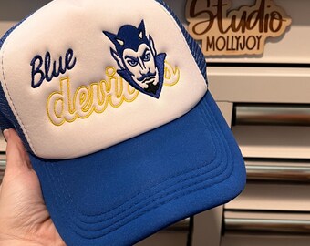 Puede incluir: Una gorra de camionero azul y blanca con un gráfico bordado de una mascota de diablo. El texto Blue está bordado en hilo azul y la palabra devils en hilo amarillo. La gorra tiene una visera azul sólida y un panel trasero de malla.