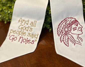Puede incluir: Una estola de graduación blanca con texto bordado en dorado que dice "And all God's people said 'Go Noles'" en el lado izquierdo y una ilustración bordada en granate de un perfil con un tocado de plumas en el lado derecho.