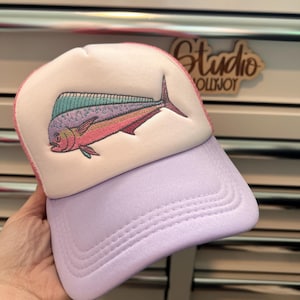 May include: A trucker hat featuring a white foam front panel with an embroidered mahi-mahi fish in shades of pink, teal, and purple. The hat has a light purple brim and matching mesh back panels. The embroidery depicts the fish in profile.