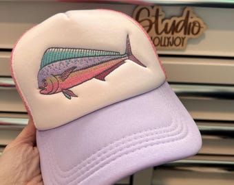 Puede incluir: Una gorra de camionero con panel frontal de espuma blanca y un bordado de un pez mahi-mahi en tonos rosa, turquesa y morado. La gorra tiene una visera de color morado claro y paneles traseros de malla a juego. El bordado muestra al pez.