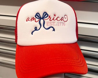 Puede incluir: Gorra de camionero roja y blanca. El panel frontal blanco tiene bordado "America" en letras rojas y rosas, un lazo azul y el texto "EST. 1776". La visera es roja y la parte trasera es de malla roja.