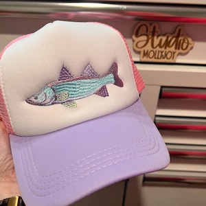 May include: A trucker hat featuring a white foam front panel with an embroidered fish in shades of teal, purple, and yellow. The hat has a solid lavender brim and pink mesh back panels. The design includes a structured crown and a curved visor.
