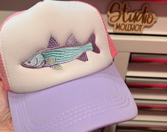 Puede incluir: Una gorra tipo trucker con panel frontal de espuma blanca y un pez bordado en tonos turquesa, morado y amarillo. La gorra cuenta con una visera de color lavanda sólido y paneles traseros de malla rosa con una corona estructurada.