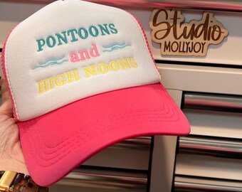 Puede incluir: Una gorra de camionero blanca y rosa intenso con el texto bordado PONTOONS and HIGH NOONS en hilo color verde azulado, rosa y amarillo. El diseño incluye dos pequeños iconos de olas azul claro a los lados de la palabra and, con malla y frente de espuma.