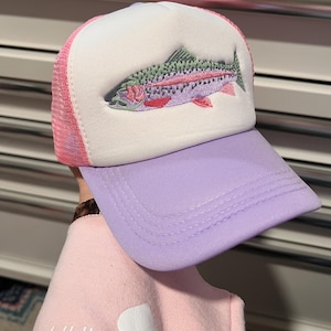 May include: A trucker hat with a white front panel, a pink mesh back, and a lavender brim. The hat features an embroidered rainbow trout design in shades of green, pink, and purple. A casual accessory for outdoor enthusiasts.