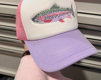 Puede incluir: Gorra tipo trucker con panel frontal blanco, parte trasera de malla rosa y visera lavanda. La gorra presenta un diseño bordado de trucha arcoíris en tonos verdes, rosas y morados. Un accesorio casual.