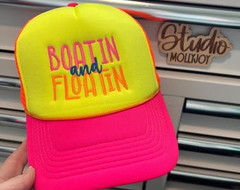 Puede incluir: Una gorra de camionero de color amarillo neón y rosa intenso con el texto bordado BOATIN and FLOATIN en hilo rosa, naranja y azul. La gorra tiene un panel frontal amarillo brillante, una visera rosa intenso y un cordón amarillo fino.