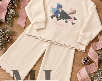 Puede incluir: Un conjunto de sudadera de manga larga y pantalón ancho color crema con bordes festoneados. La sudadera presenta un gráfico en punto de cruz de dos figuras cargando un árbol de Navidad y el nombre Ruthie bordado en color rosa.