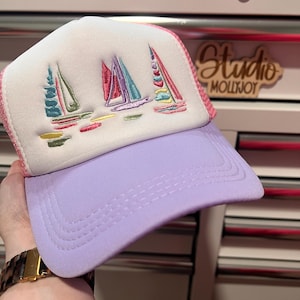 May include: A trucker hat with a white foam front panel featuring an embroidered design of three sailboats in shades of pink, teal, and yellow. The hat has a solid lavender brim and a matching lavender mesh back panel with pink trim detailing.