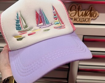 Puede incluir: Una gorra tipo trucker con panel frontal de espuma blanca que presenta un diseño bordado de tres veleros en tonos rosa, turquesa y amarillo. La gorra tiene una visera de color lavanda y un panel trasero de malla a juego en lavanda.