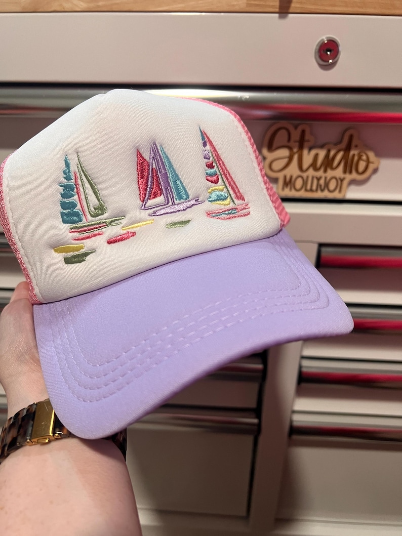 May include: A trucker hat with a white foam front panel featuring an embroidered design of three sailboats in shades of pink, teal, and yellow. The hat has a solid lavender brim and a matching lavender mesh back panel with pink trim detailing.
