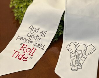 Puede incluir: Una estola de graduación blanca con texto bordado en negro que dice And all God's people said, Roll Tide, con la palabra Roll Tide en rojo. El otro lado de la estola presenta el contorno bordado en negro de un elefante mirando al frente.