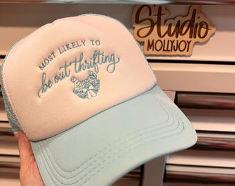 Puede incluir: Una gorra de camionero azul claro y blanca con el texto bordado "MOST LIKELY TO be out thrifting" en hilo azul, acompañado de un pequeño gráfico bordado en azul de un pollo con un sombrero de fiesta y un lazo.