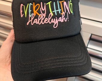 Puede incluir: Gorra de camionero negra con el texto bordado "EVERYTHING Hallelujah!" en hilo colorido. La gorra tiene un panel frontal y una visera negros, con una parte trasera de malla blanca. El texto está en una fuente multicolor juguetona.