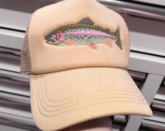 Puede incluir: Gorra de camionero color melocotón con parte trasera de malla y visera curva. La gorra presenta un diseño bordado de trucha arcoíris en tonos rosa, verde y gris. Un accesorio clásico para los amantes del aire libre.