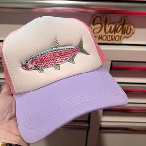 May include: A trucker hat featuring a white foam front panel with an embroidered pink and green fish graphic. The hat has a light purple brim and a pink mesh back. The front panel displays a detailed fish design with scales in shades of pink.