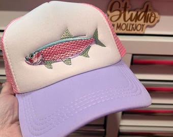 Puede incluir: Una gorra de camionero con panel frontal de espuma blanca y un gráfico bordado de un pez rosa y verde. La gorra tiene una visera de color violeta claro y una parte trasera de malla rosa. El diseño del pez muestra escamas en tonos rosa.