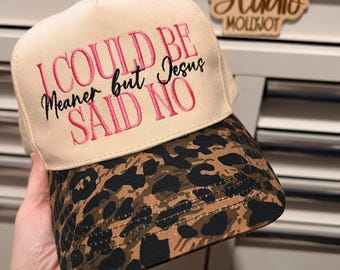 Puede incluir: Una gorra de béisbol beige con visera de estampado de leopardo. El panel frontal presenta texto bordado en rosa y negro que dice: I COULD BE Meaner but Jesus SAID NO. La gorra tiene una corona estructurada y una visera curva con costuras.