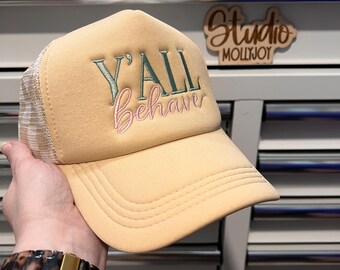 Puede incluir: Una gorra tipo camionero de color melocotón con una parte trasera de malla blanca y un diseño bordado en el panel frontal. El texto dice Y'ALL en letras mayúsculas verde oscuro con la palabra Behave bordada debajo en cursiva rosa.