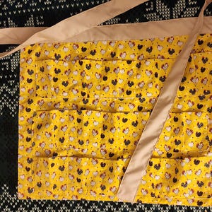 May include: A yellow egg gathering apron with a repeating pattern of black and white chickens. The apron has multiple pockets and a tan strap that goes around the neck and waist. The apron is made of fabric.