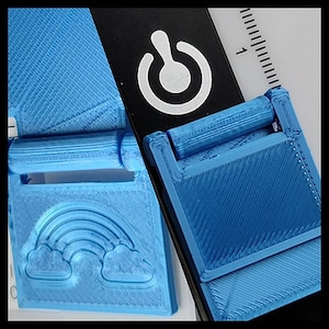 Op de afbeelding: Een blauwe plastic gesp met een regenboog- en wolkenontwerp. De gesp is bevestigd aan een zwarte band met een wit stroomknopsymbool.