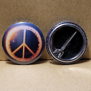 May include: A round button featuring a peace sign design. The peace sign is a dark orange colour and is surrounded by a dark blue background. The reverse of the button is silver with a pin.