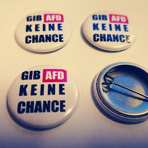 Może przedstawiać: Trzy białe okrągłe guziki z czarnym i różowym wzorem. Guziki mają napis "Gib AfD Keine Chance". Jeden guzik jest otwarty, pokazując metalowe plecy i szpilkę.