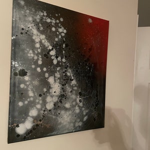 May include: Abstract painting on canvas with black, white, and red paint splatters. The painting has a galaxy-like appearance with white and black splatters on a dark background, transitioning to a red background on the right side.