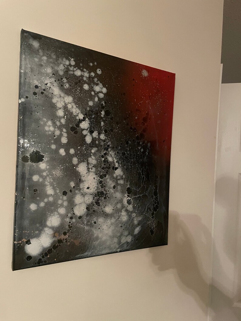 May include: Abstract painting on canvas with black, white, and red paint splatters. The painting has a galaxy-like appearance with white and black splatters on a dark background, transitioning to a red background on the right side.
