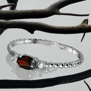 May include: A silver ring with a red oval gemstone and two smaller clear gemstones. The ring is beaded and has the inscription "JEWELETS" on the band.