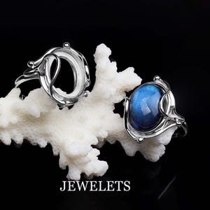 May include: Two silver rings with oval-shaped settings. One ring has a blue gemstone, the other is empty. The rings are displayed on a white coral-like surface.