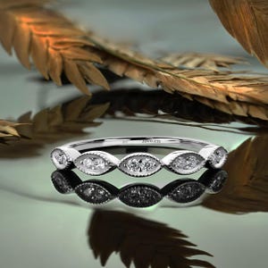 Op de afbeelding: Een zilveren ring met een reeks marquise-vormige zettingen, elk met kleine, heldere edelstenen. De ring ligt op een reflecterend oppervlak, met het woord "JEWELETS" op de band.