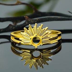 May include: A gold-colored ring with a sunburst design. The ring features a central circular setting surrounded by radiating lines, creating a sun-like appearance. The band is a double-layered design. The ring is highly reflective.