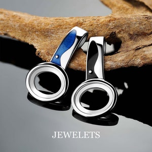 May include: Two silver pendant charms with a circular opening. One charm has a blue enamel design and the other has a black enamel design. The charms are on a wooden surface.