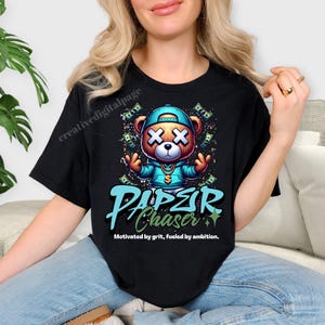 Może przedstawiać: Czarny t-shirt z kresk&oacute;wkowym misiem w niebieskiej czapce i bluzie z kapturem, z symbolami dolara i napisem "PAPER CHASER". Miś ma X na każdym oku. Tekst "Motywowany odwagą, napędzany ambicją" znajduje się poniżej.