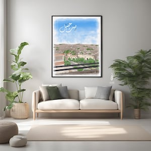 Puede incluir: Una impresión artística enmarcada que presenta un paisaje con un cielo azul y un paisaje urbano. La obra de arte incluye caligrafía árabe en la parte superior, con el texto en inglés "TINA JBEIL" debajo. La impresión se muestra encima de un sofá beige.