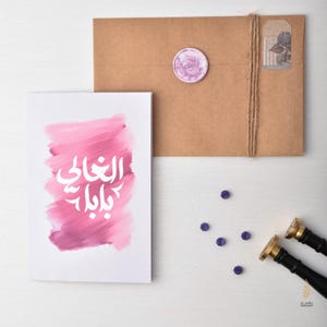 以下が含まれることがあります： ピンクの水彩画の背景と白いアラビア語のテキスト「世界で最高の」が書かれた白いカード。カードは、茶色の封筒と紫色のワックスシールのある白い表面の上に置かれています。