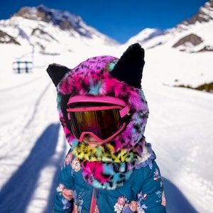 May include: A child wearing a vibrant pink and purple leopard print ski hood with black ears and pink ski goggles. The child is wearing a blue floral jacket, standing on a snowy ski slope with mountains in the background.
