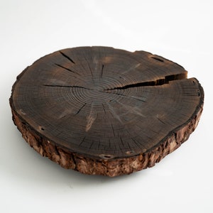 Puede incluir: Una rodaja de madera redonda con un borde de corteza natural. La madera es de color marrón oscuro y tiene un patrón de veta visible. El centro de la rodaja tiene un patrón circular de anillos.