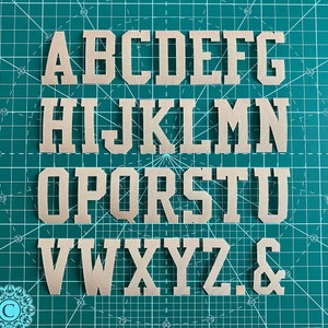 May include: A complete alphabet of beige felt letters, including the ampersand symbol, displayed on a green cutting mat. The letters are in a bold, block style, suitable for crafting or customisation. The mat features grid lines for accurate alignment.