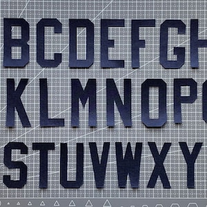May include: A full alphabet of navy blue felt letters, cut in a blocky, sans-serif font. The letters are arranged in three rows on a gray cutting mat with grid lines. The letters are evenly spaced and clearly defined.