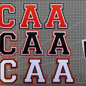 Peut inclure: Ensemble de lettres en tissu, dont les lettres "C" et "A", en rouge, noir et blanc avec des bordures contrastées. Les lettres sont disposées sur un tapis de coupe gris, avec des échantillons de tissu.