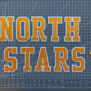 Peut inclure: Le texte NORTH STARS est affiché dans une police grasse de style bloc avec des lettres orange soulignées de blanc. Les lettres sont placées sur un tapis de découpe gris foncé avec une grille et des repères de mesure en centimètres.