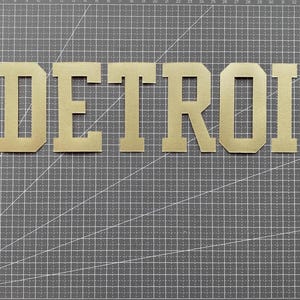 Peut inclure: Lettres dorées formant le mot DETROIT dans une police en bloc, placées à côté d'une règle en métal noir indiquant une hauteur d'environ 15 cm pour les lettres, sur un tapis de découpe quadrillé gris foncé avec des marquages blancs.