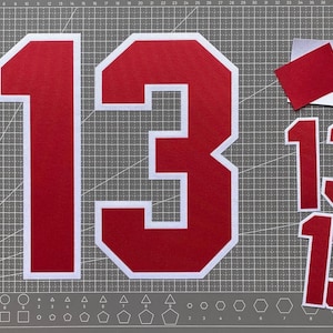 May include: A large red number 13 with a thick white border, accompanied by two smaller red number 13 cutouts with white borders and a small red rectangular scrap, all displayed on a gray cutting mat with white grid lines and geometric shapes.