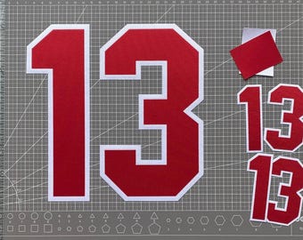 May include: A large red number 13 with a thick white border, accompanied by two smaller red number 13 cutouts with white borders and a small red rectangular scrap, all displayed on a gray cutting mat with white grid lines and geometric shapes.