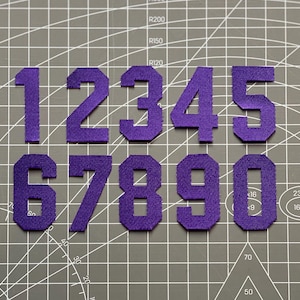 May include: A collection of purple fabric numbers, ranging from 1 to 0, arranged on a gray cutting mat. The numbers are in a bold, blocky font, and appear to be iron-on patches or appliques. The background has grid lines.