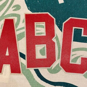 Peut inclure: Gros plan sur un motif de tissu avec les lettres "ABC" en rouge bordées de blanc. Le fond est crème avec des motifs abstraits verts et turquoise, créant une esthétique vintage.