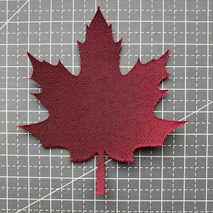 May include: A deep red maple leaf cutout made of a woven fabric material. The leaf features a distinct five-pointed shape with serrated edges and a central stem at the bottom, displaying a clear, uniform grid-like texture across its surface.